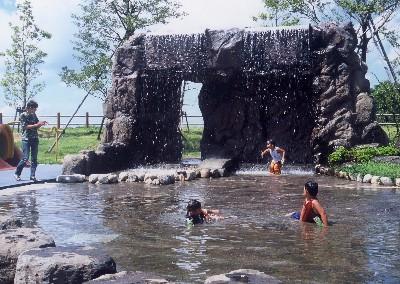 岩から滝のように水が落ちる公園内の水場で遊ぶ子どもたちの写真