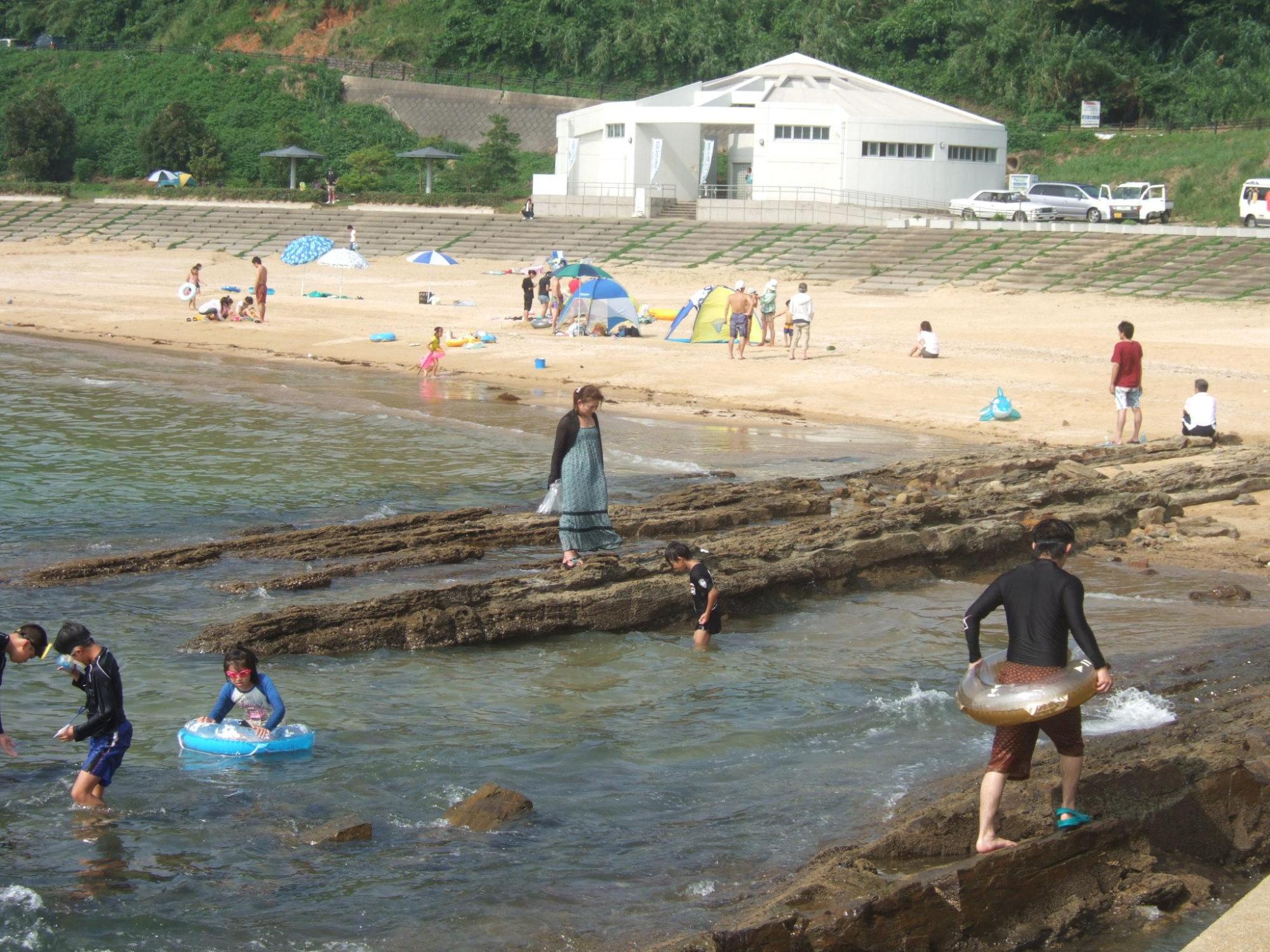 背景には白いトイレやシャワーの施設、海水浴場では浮き輪を持って遊ぶ子どもたちや浜辺のパラソルの写真